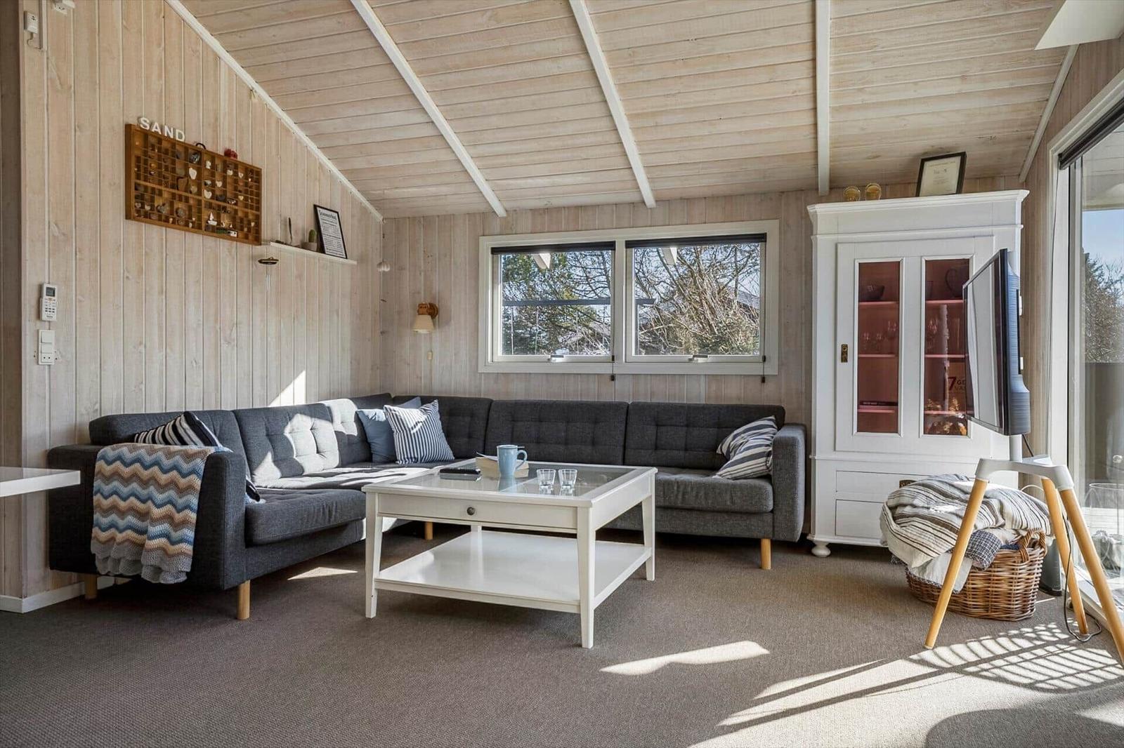Living room with gray sofa, white table, and wood walls. Windows and cabinet are visible.