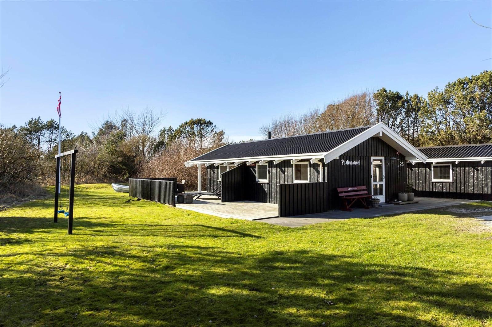 Black wooden house with terrace, garden, and swing. Flag and trees in background.