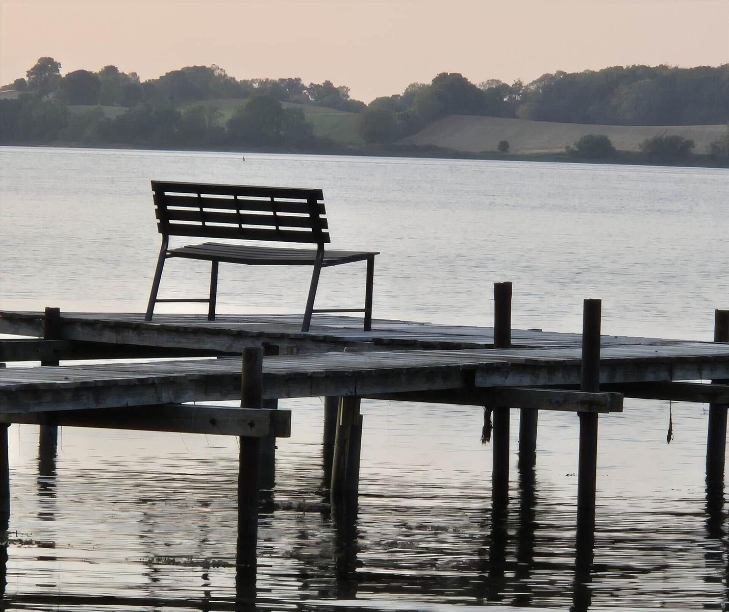 A bench sits on a wooden platform over water. Hills and trees are visible in the background.