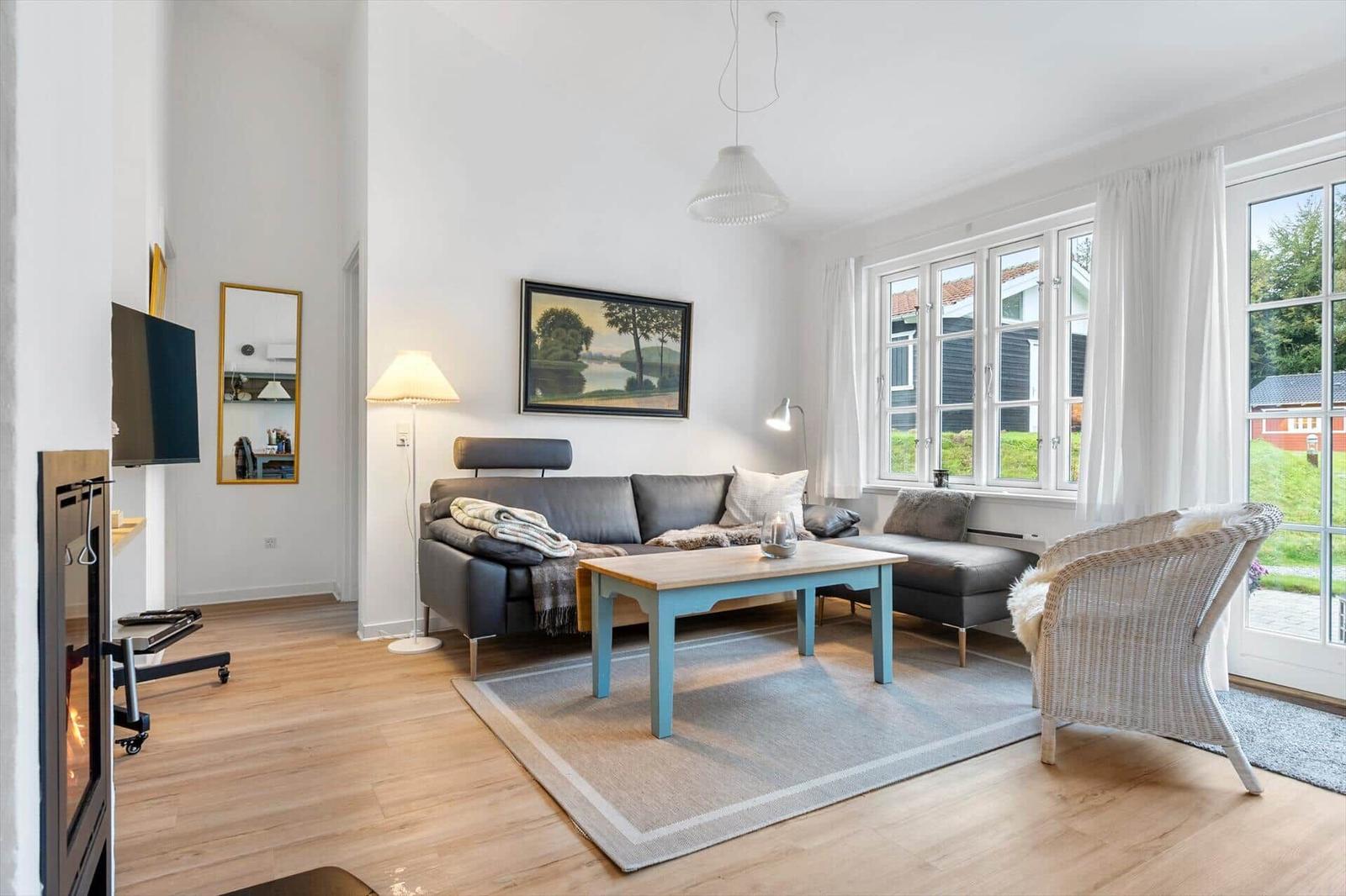 Living room with sofa, table, and chair. Windows show garden and house.