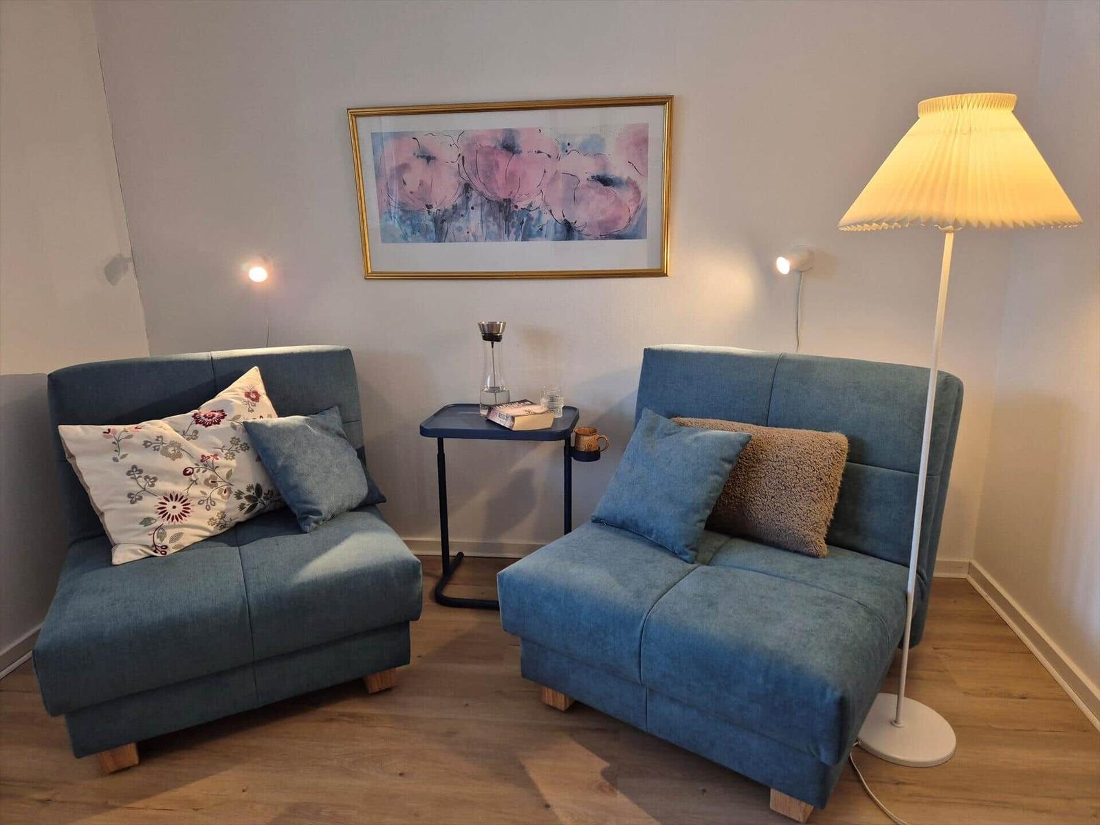 Two blue seating units with cushions, table, and floor lamp in a room with wooden floor.