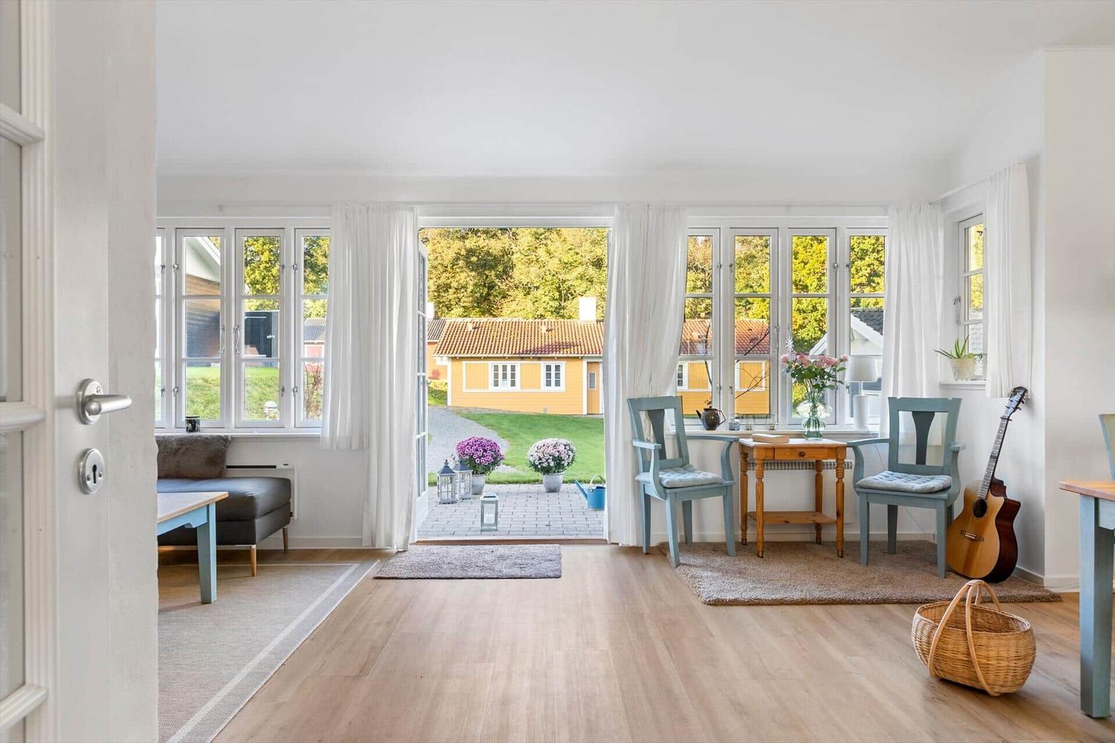 A room with wooden floor, windows, and a view of a garden. There is a table with two chairs and a guitar.