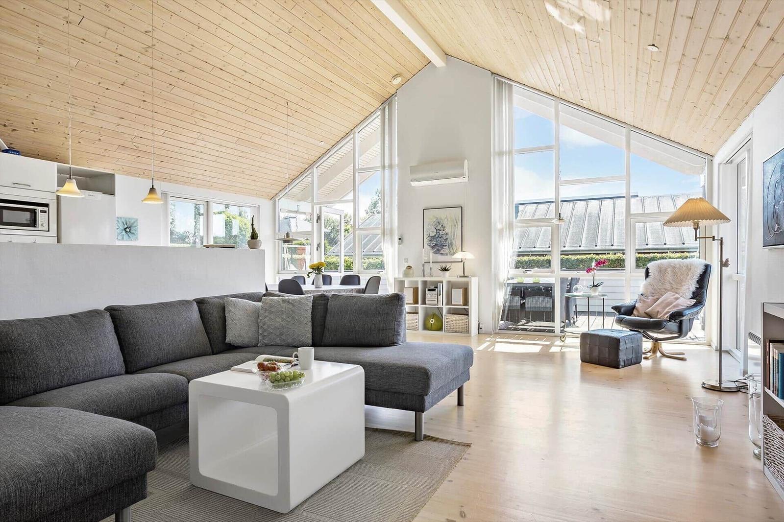Living room with gray sofa, white table, and wooden ceiling. Large windows and light.