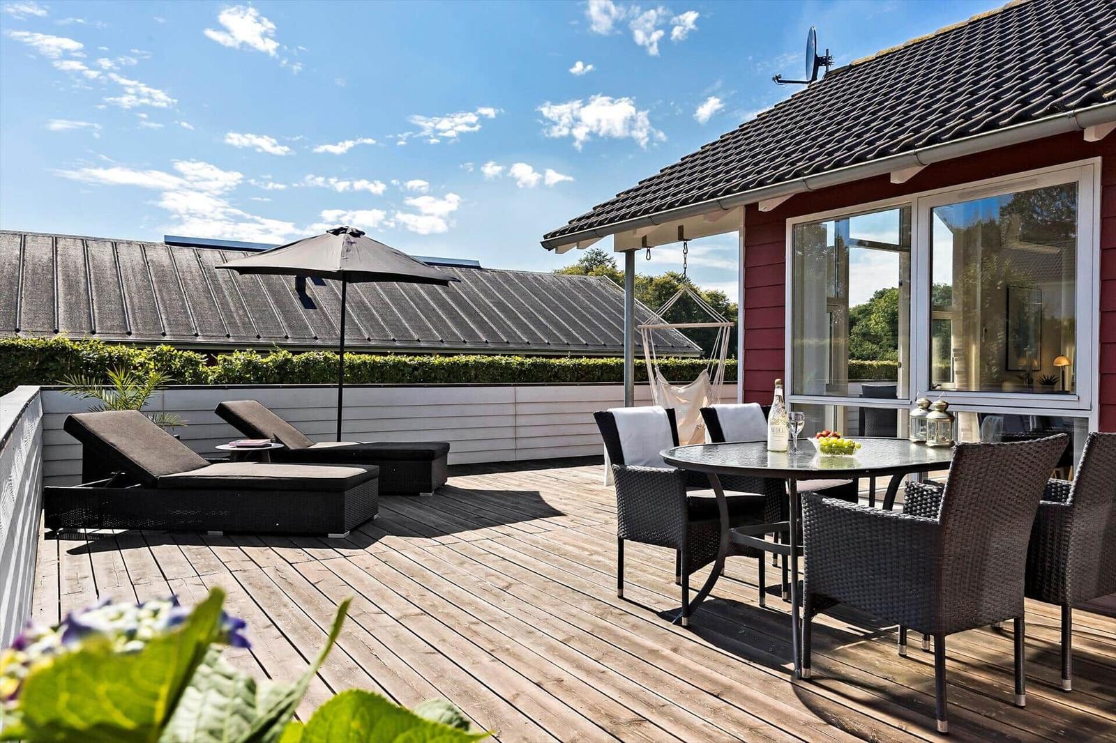 Terrace with table, chairs, loungers, and umbrella. Background: house and hedge.