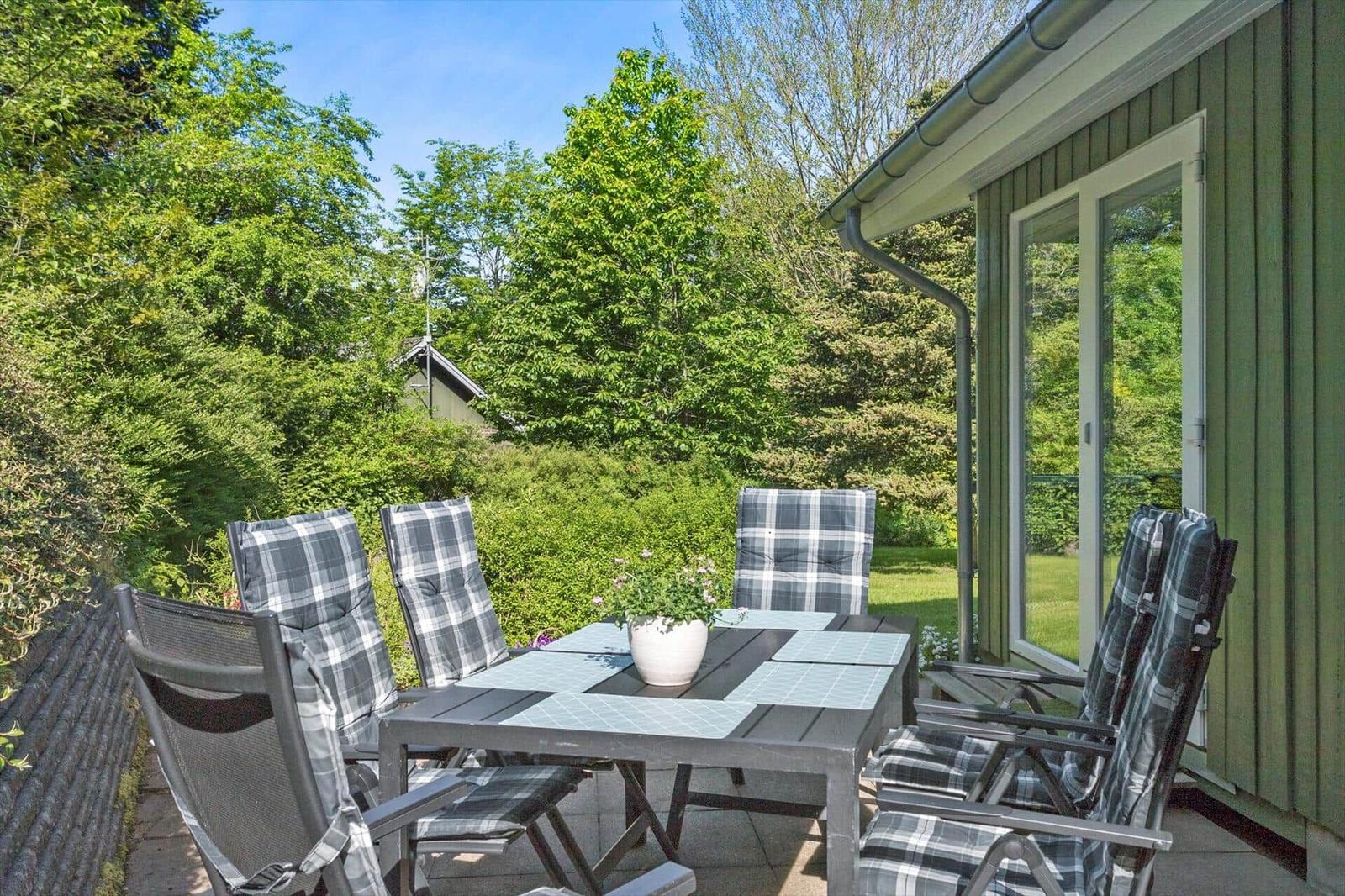 Terrace with table and chairs, surrounded by greenery and a house.