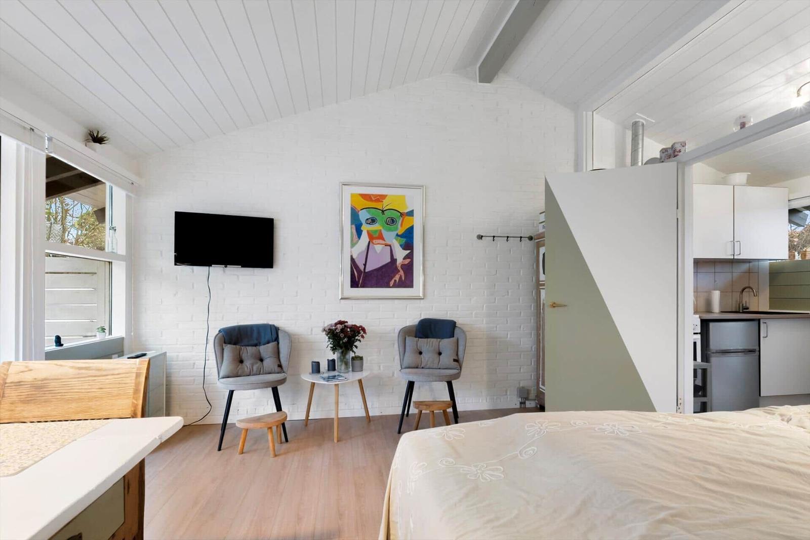 Room with bed, TV, two chairs, and window. Kitchen visible in background.