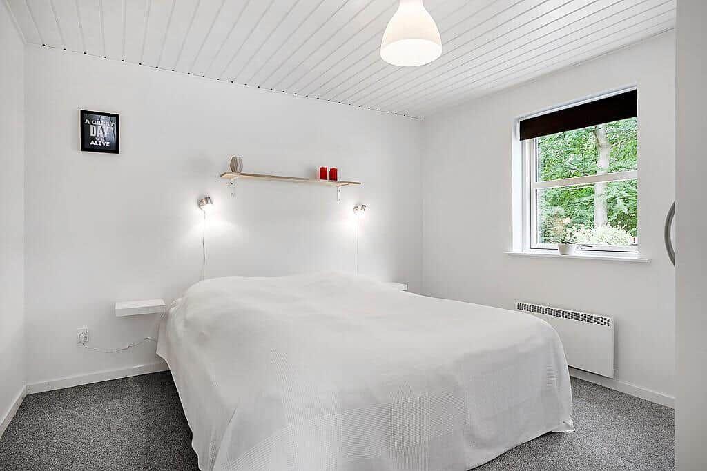 Bedroom with white bed, window, and wooden shelf.