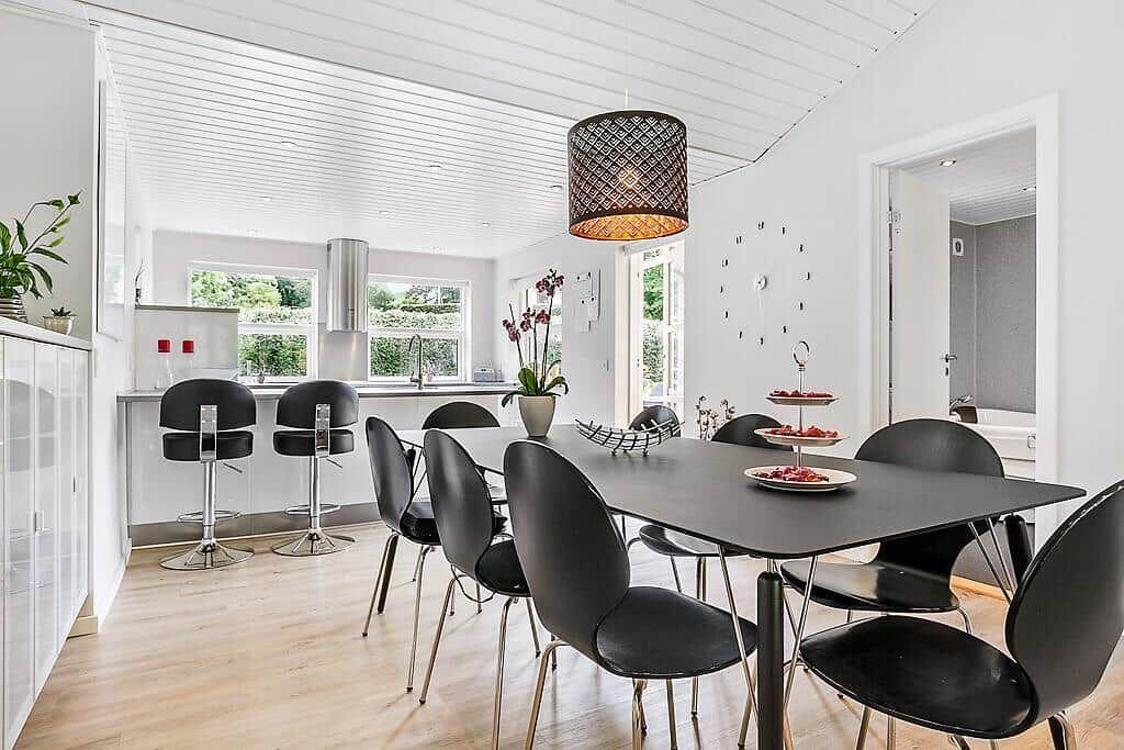Dining area with black chairs and table. Kitchen in background with windows.