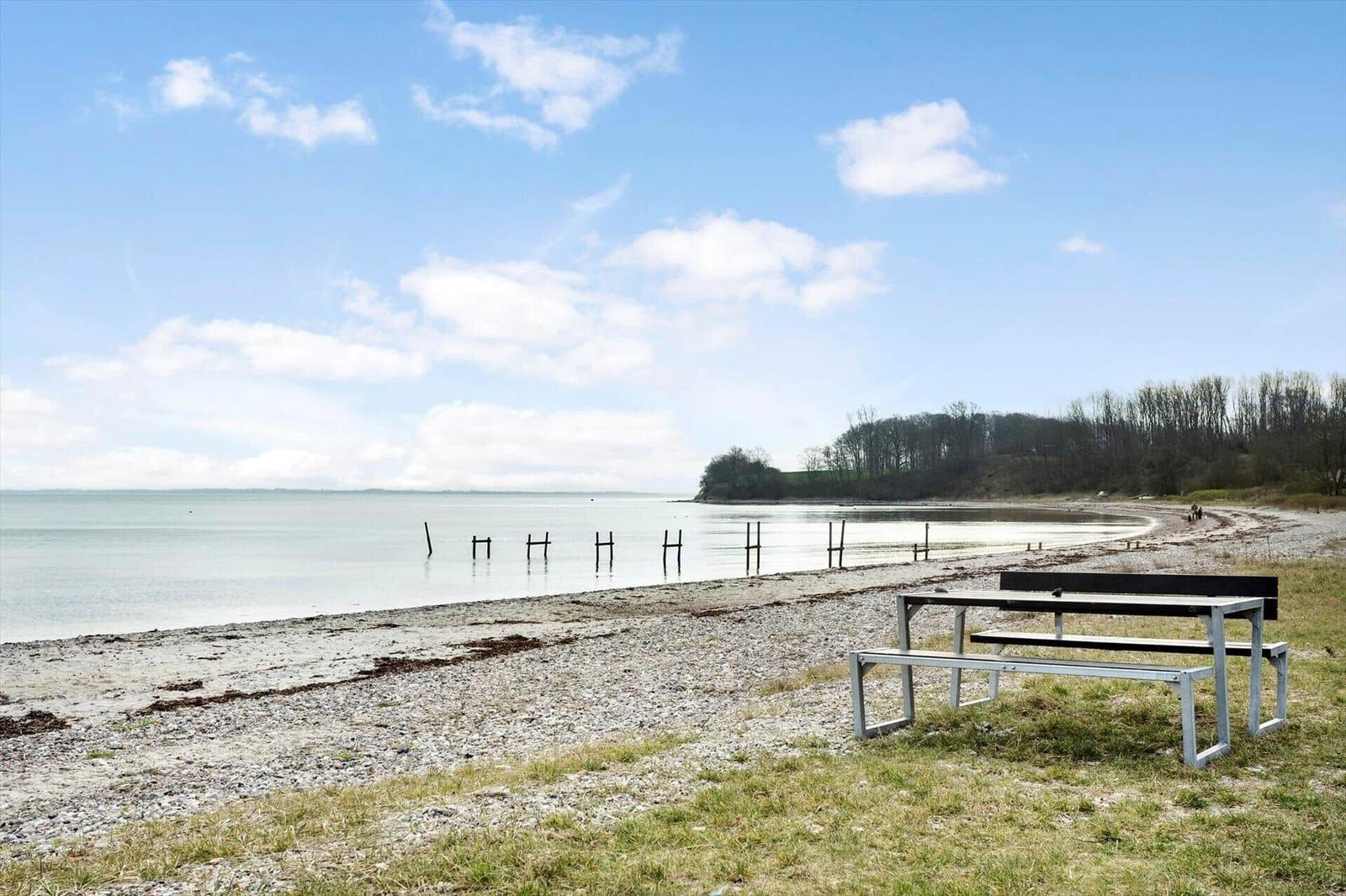 A picnic table stands by the shore next to a lake. Trees and a clear sky are visible in the background.
