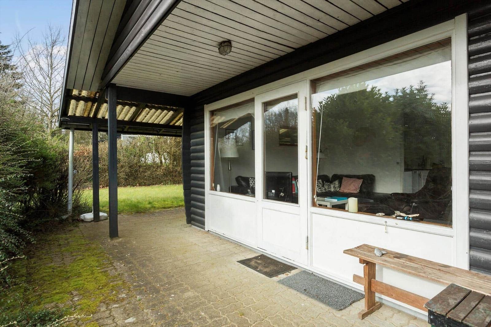 House with large window, terrace, and garden. View into living room with sofa.