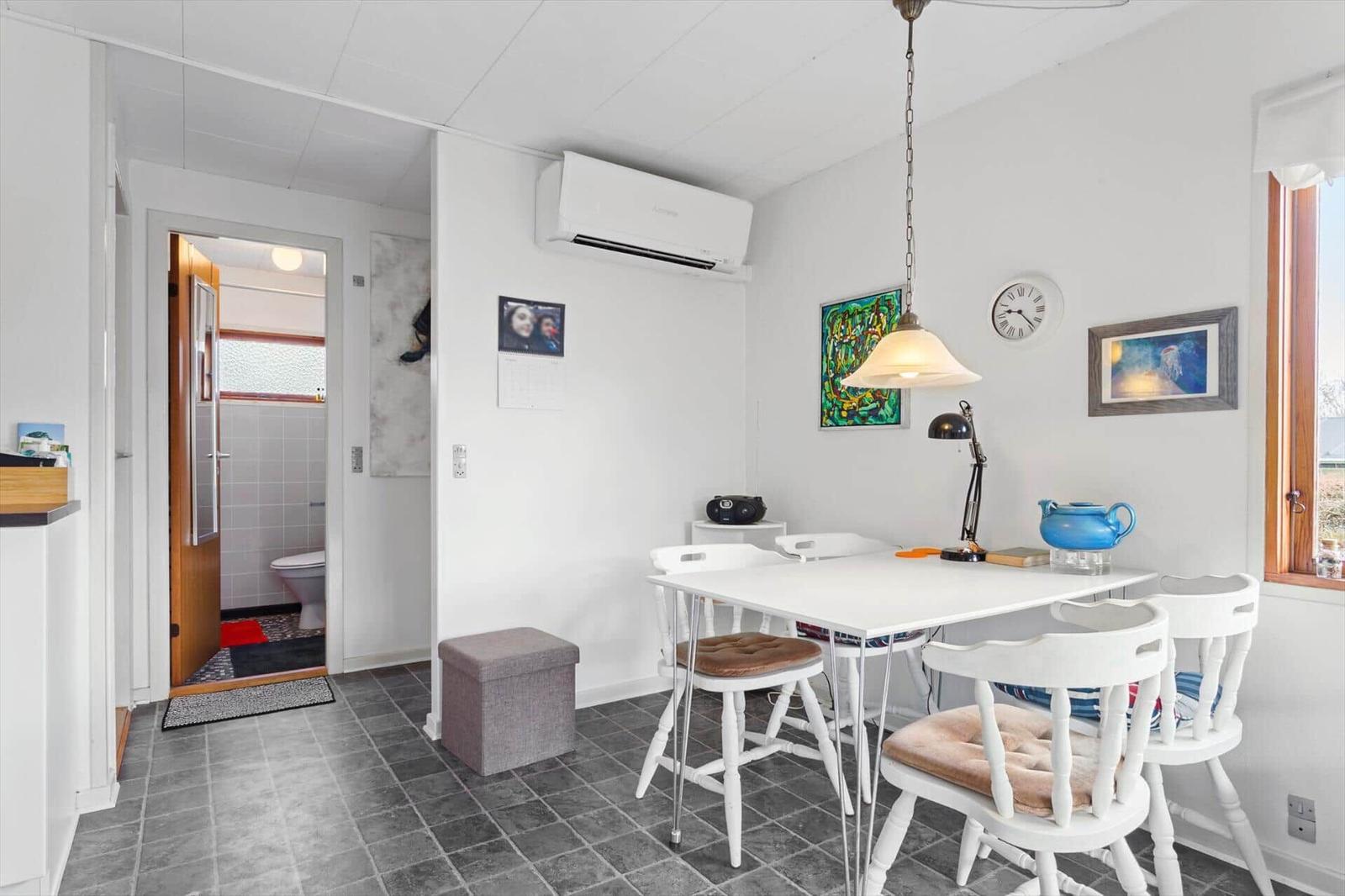 Dining area with white table and chairs, wall clock, and artwork. Passage to the toilet.
