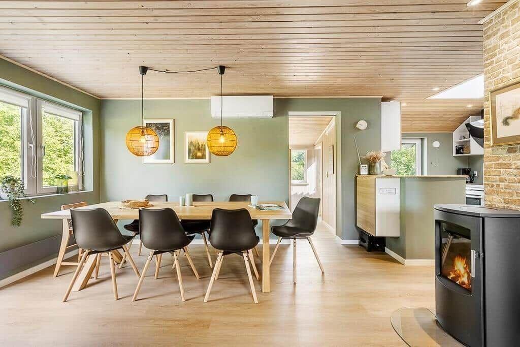 Dining area with table, chairs, and wood-burning stove. Wooden ceiling and windows with green view.