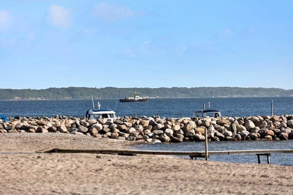 Beach with stone breakwater and boats. In the background, sea and forested shore.