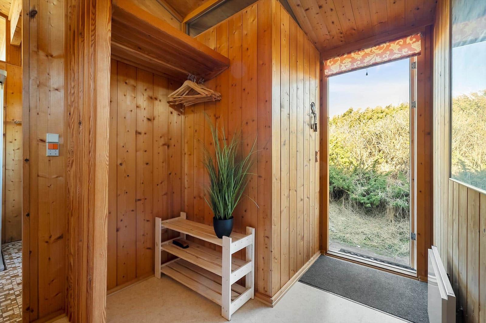 Entrance with wooden walls, shoe rack, and plant. Door opens to green outdoor view.