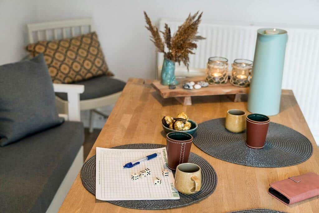 Wooden table with mugs, dice, and candles on placemats. Background: seating and dried plants.
