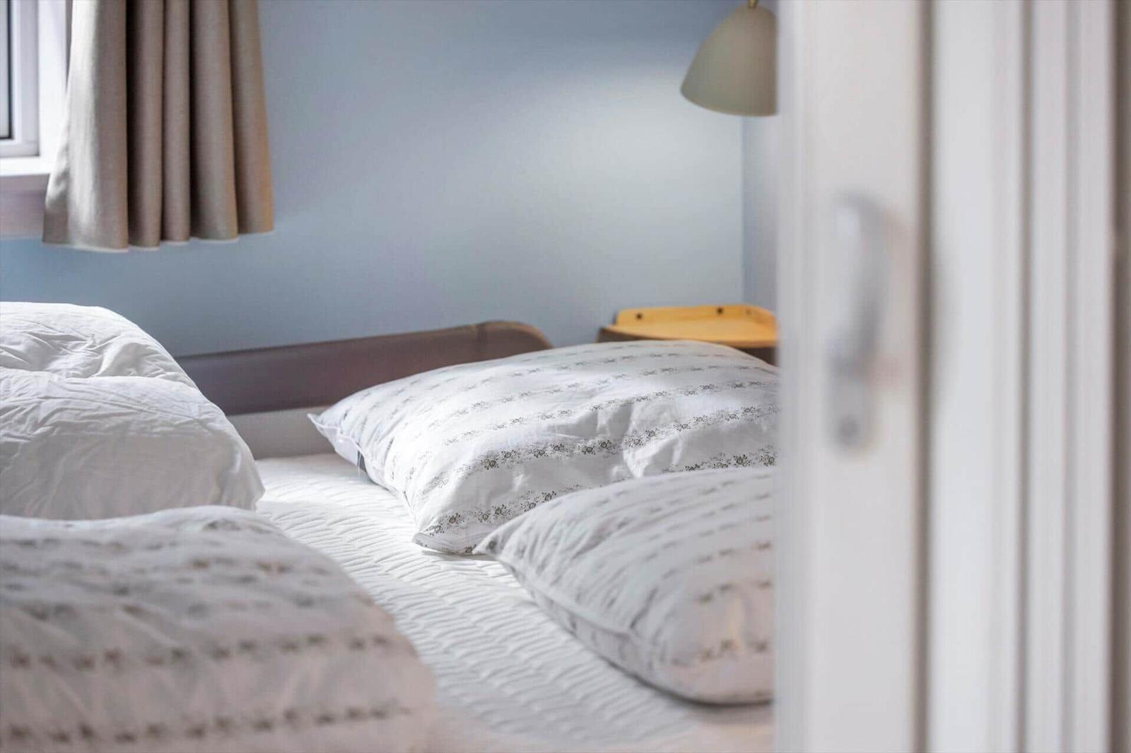 Schlafzimmer mit Bett, Kissen und Nachttisch. Vorhang und Lampe sind sichtbar.