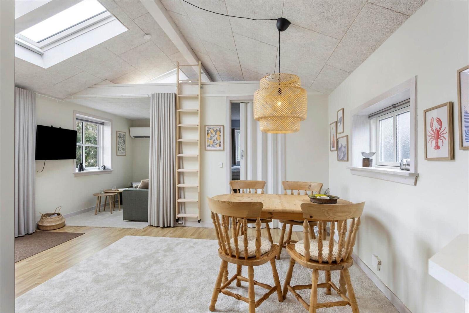 Essbereich mit Holztisch und Stühlen unter einer Deckenlampe. Im Hintergrund Wohnbereich mit Sofa und TV.