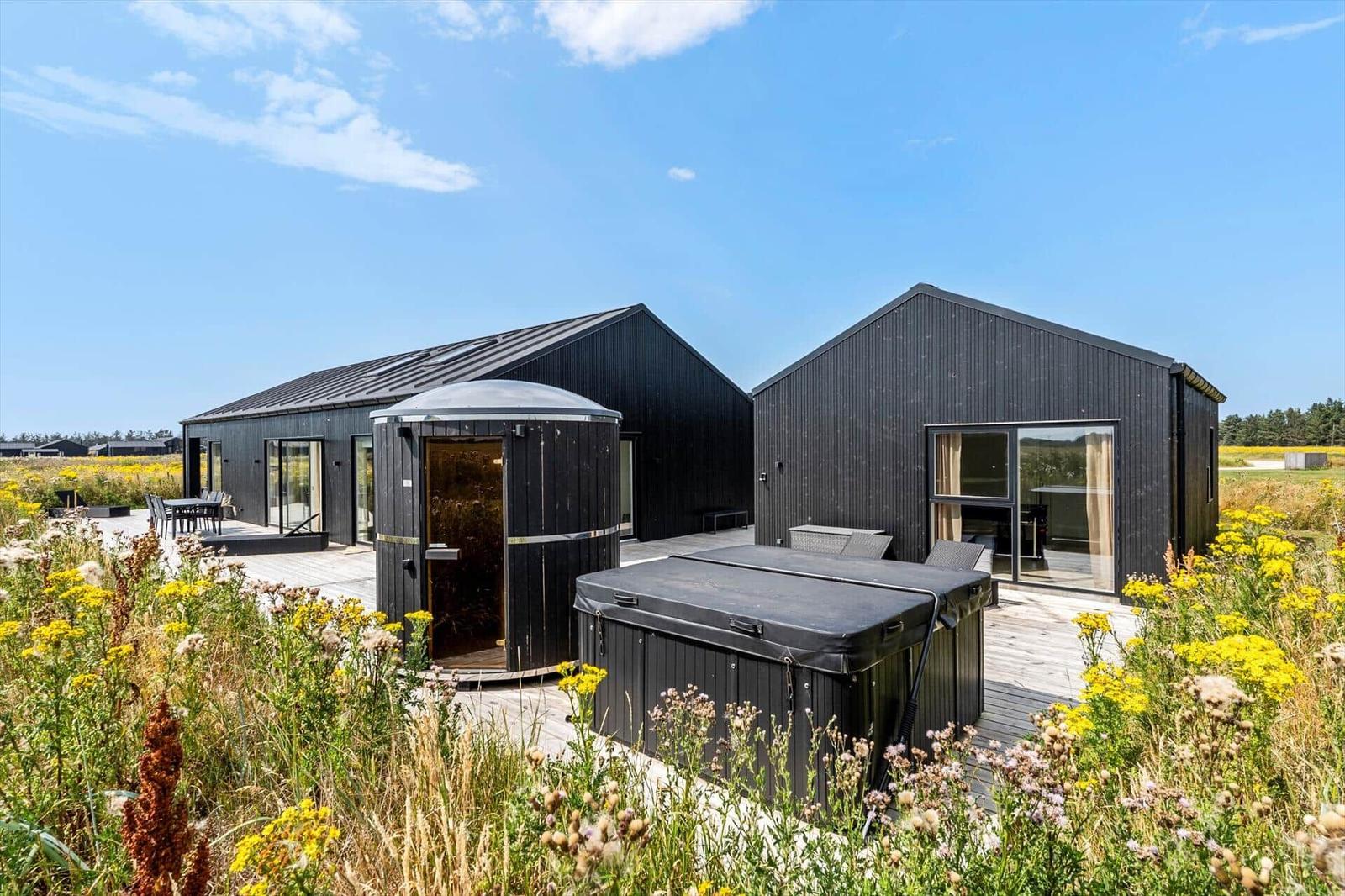 Zwei schwarze Holzhäuser mit Sauna und Whirlpool auf einer Terrasse inmitten von Blumen.