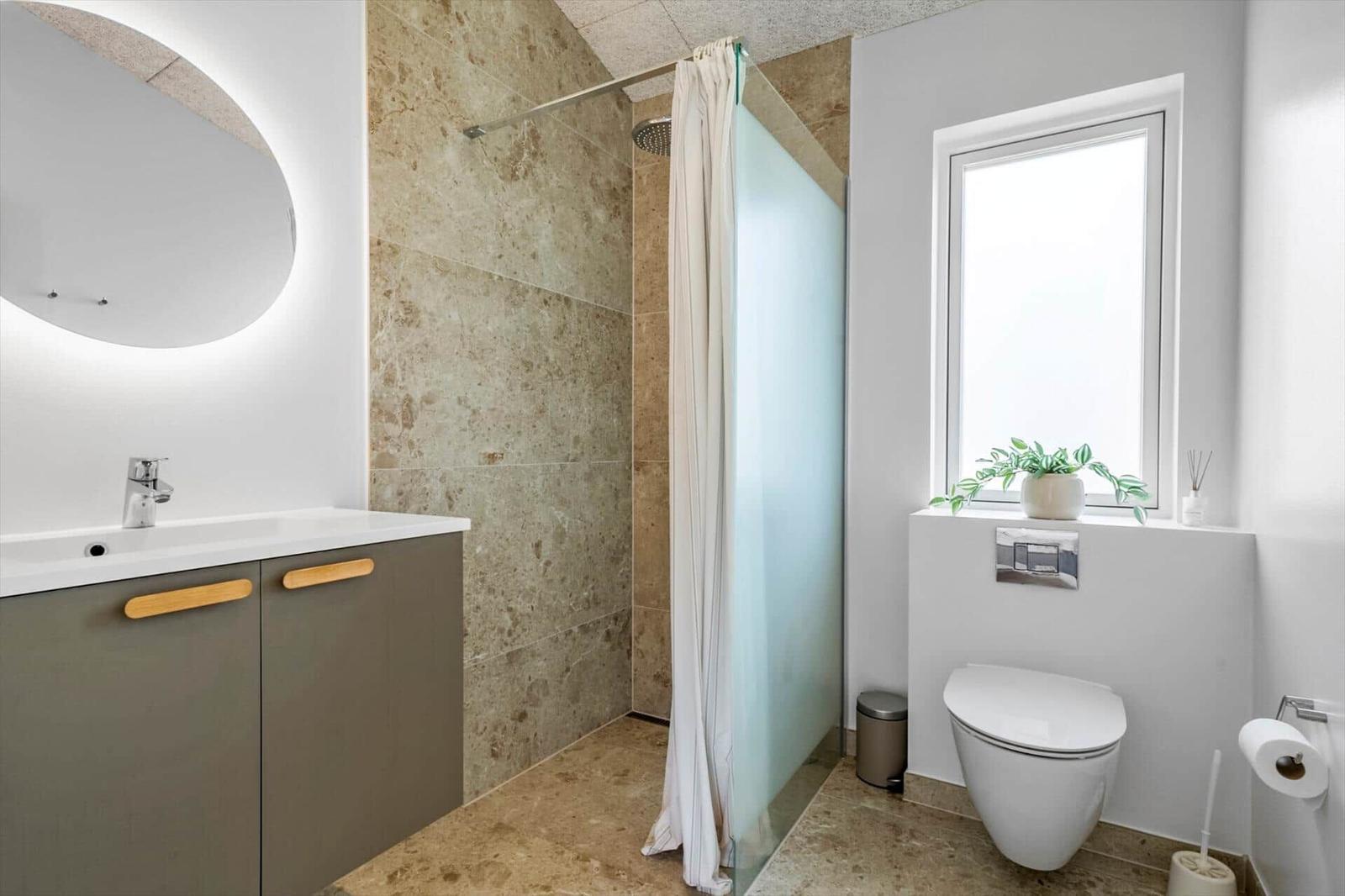 Badezimmer mit Dusche, Waschbecken und Toilette. Steinwand und Fenster sichtbar.