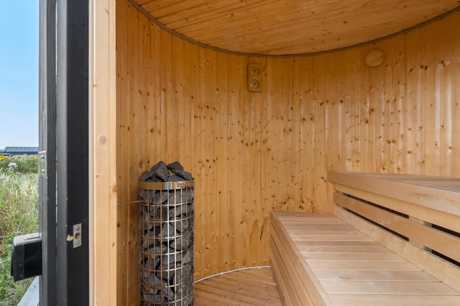 Holzsauna mit Sitzbank und Steinbehälter. Tür offen mit Blick auf Grünfläche.