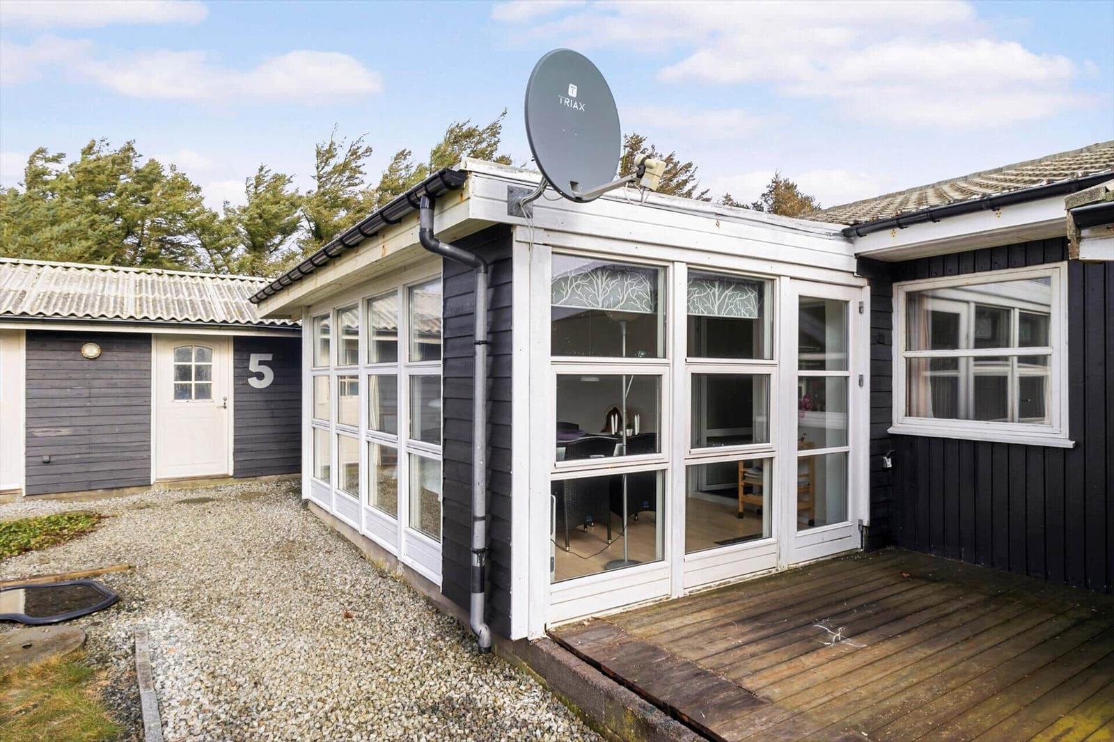 House with glass front, wooden terrace, and satellite dish. Rear yard with gravel and outbuilding.