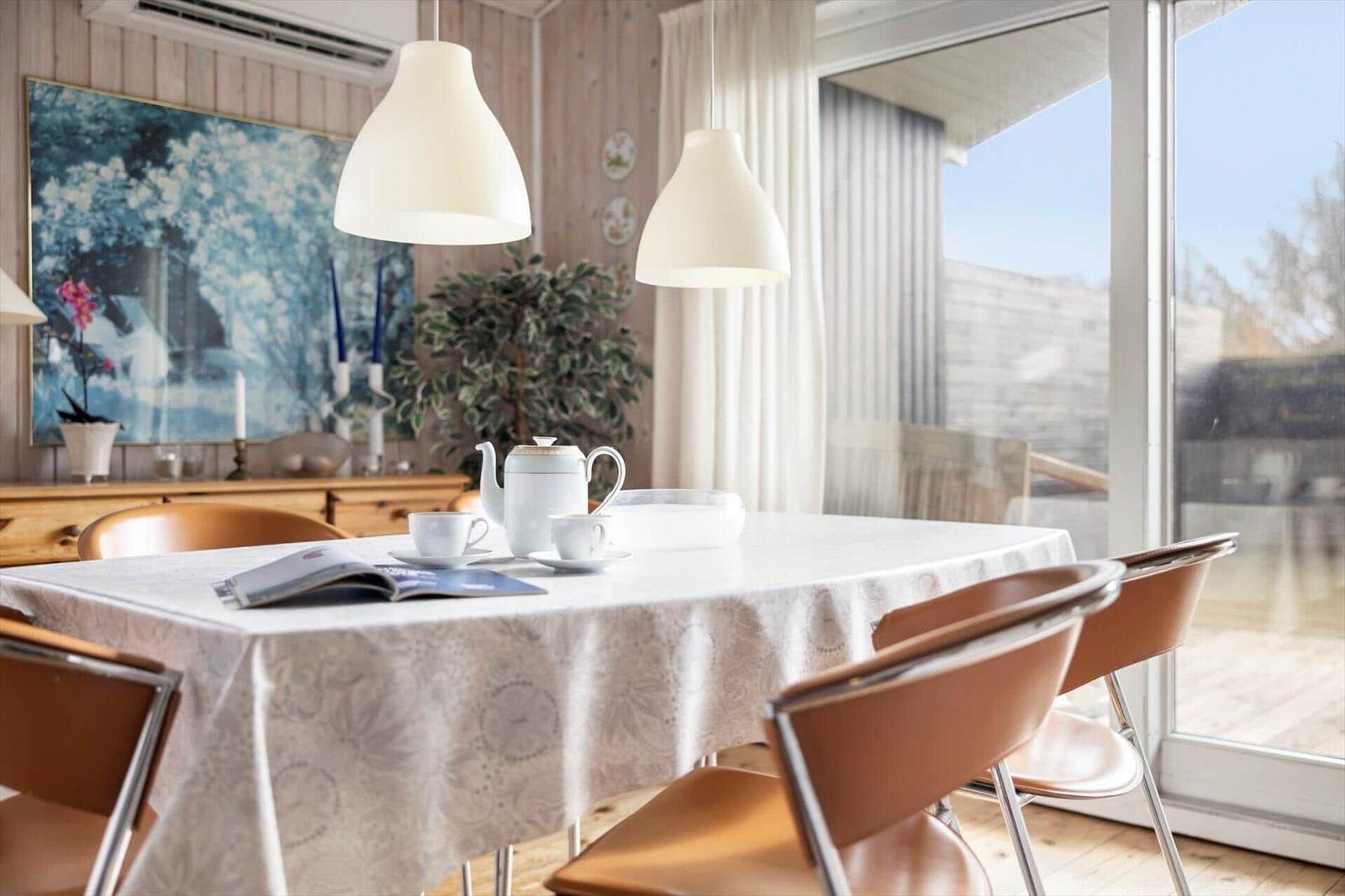 Dining table with tablecloth, cups, and teapot. Bright lamps and large windows.