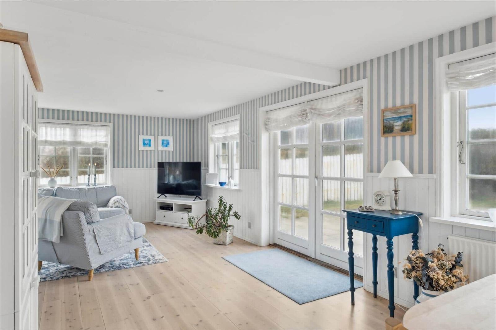 Living room with sofa, TV, and windows. Striped wallpaper and wooden floor.
