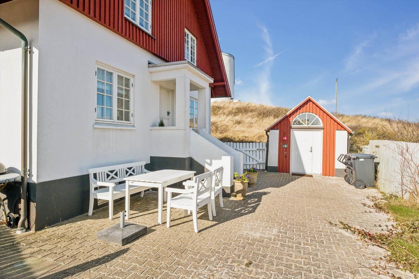 House with white seating area and red extension. Plants and pavilion in background.