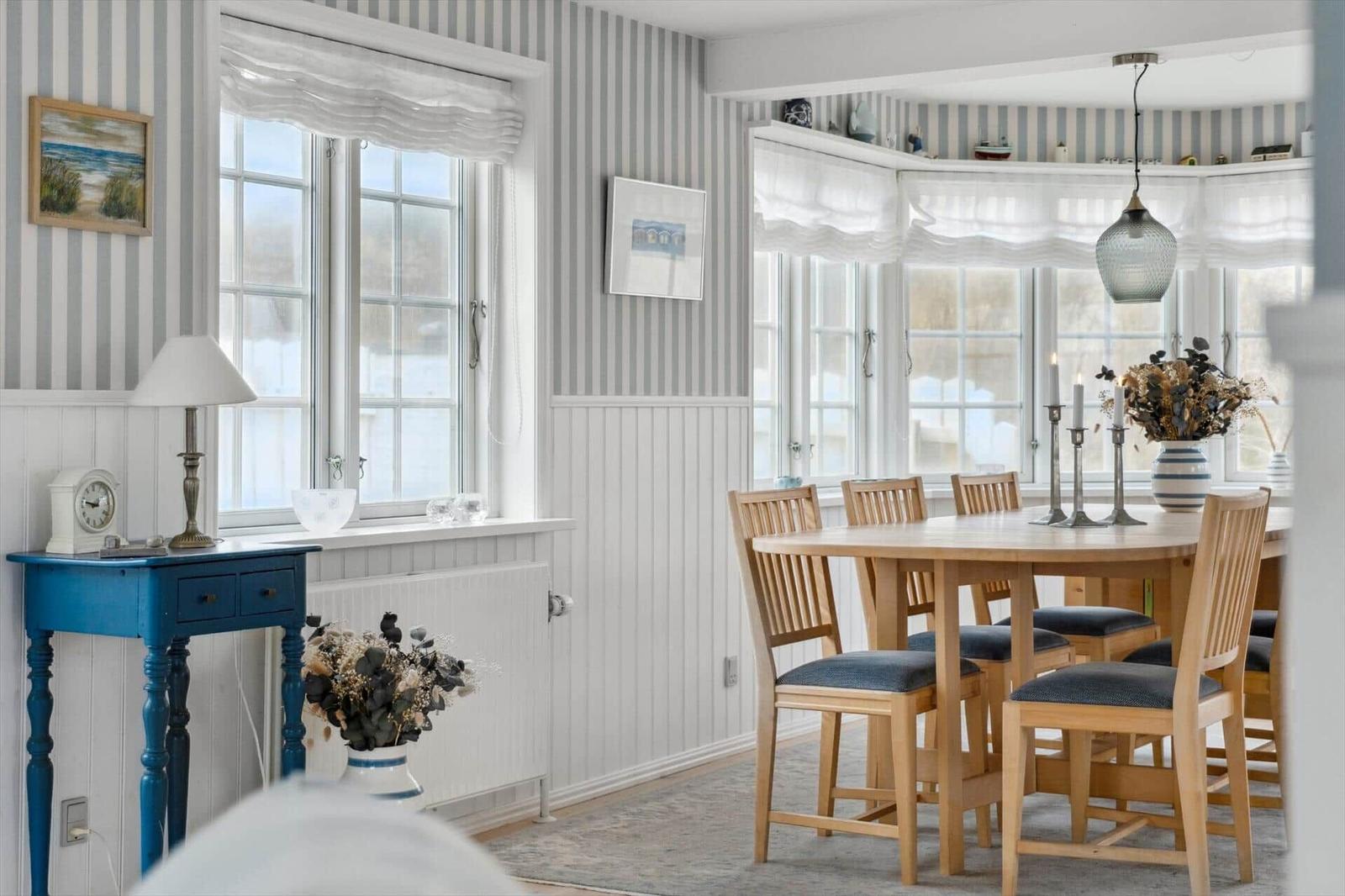 Dining room with wooden table, chairs, and windows. Walls with striped wallpaper.