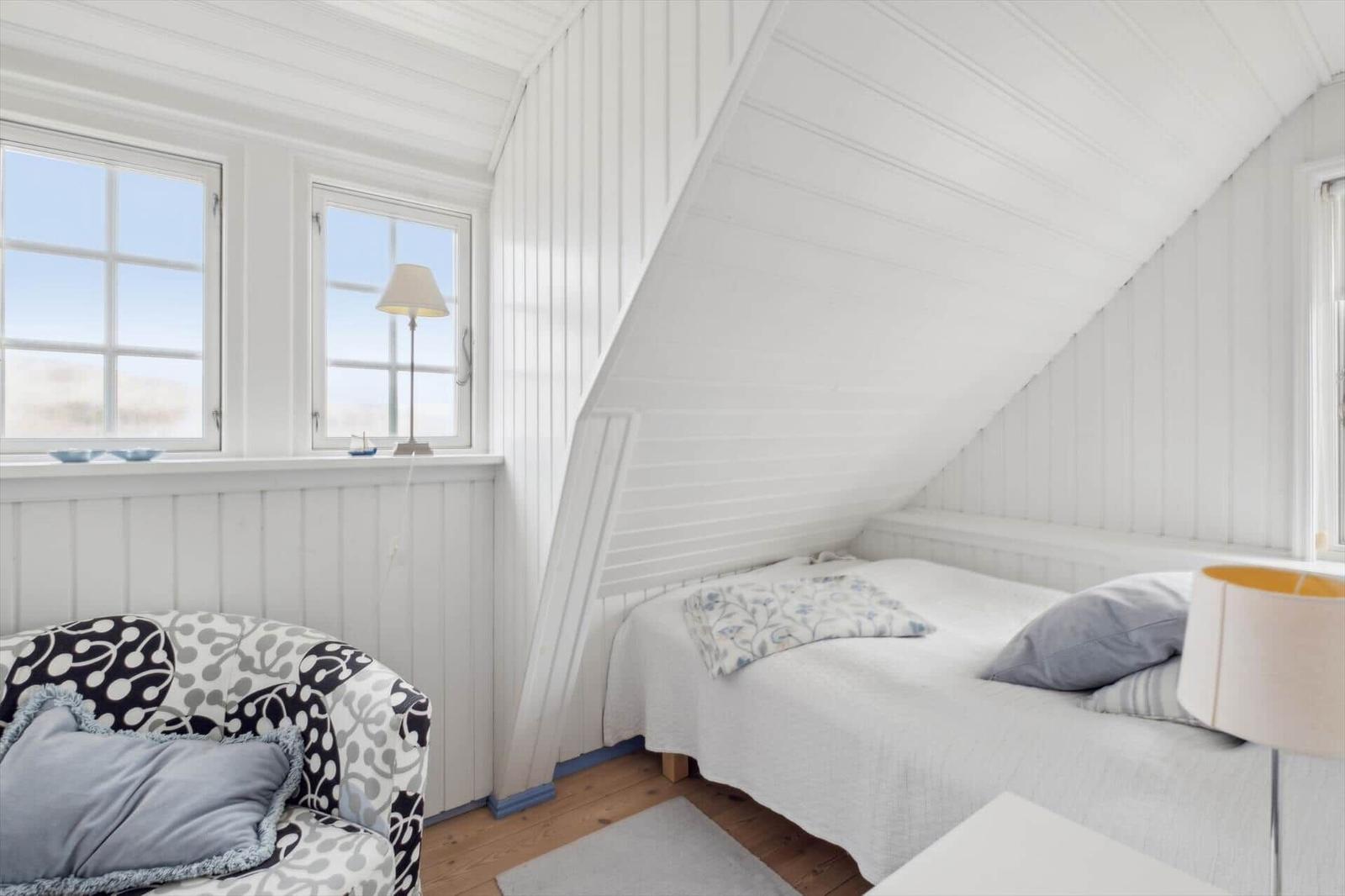 Bedroom with sloped ceiling, bed and armchair. Windows with view outside.