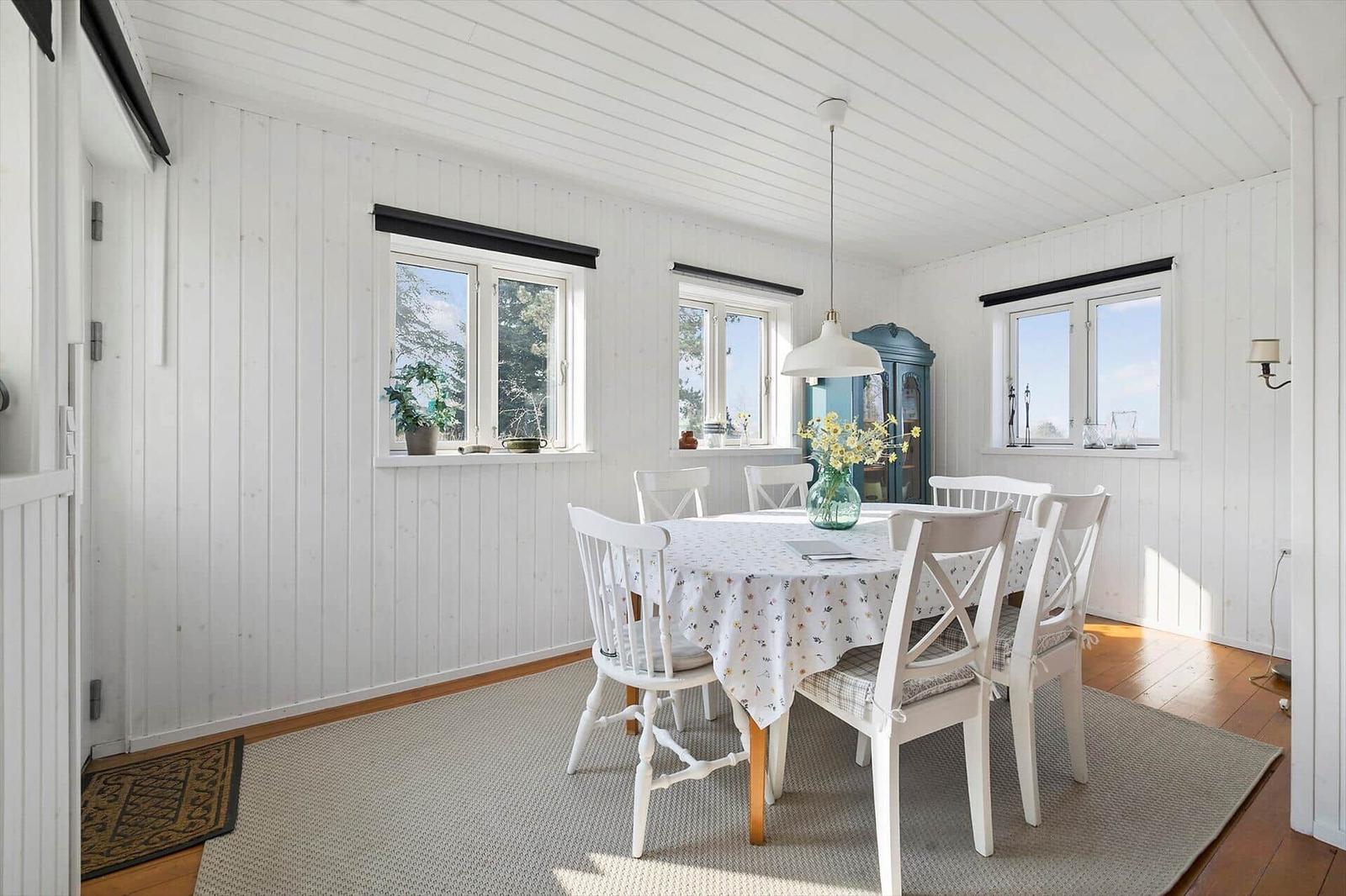 Dining room with white tablecloth and chairs, wooden floor, and windows with view outside.