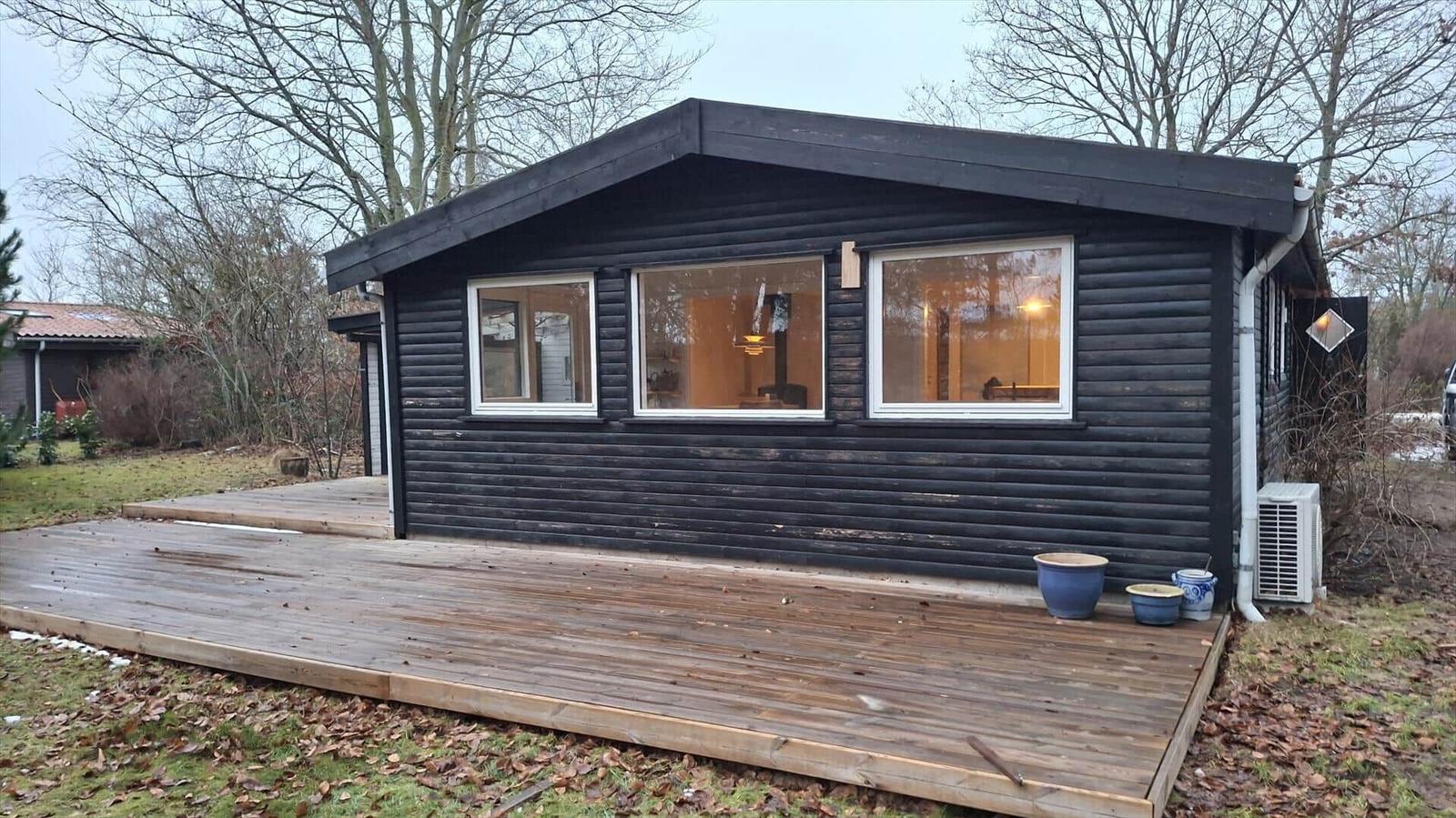 Schwarzes Holzhaus mit Holzterrasse und Fenstern. Im Inneren sind Möbel und Beleuchtung sichtbar.