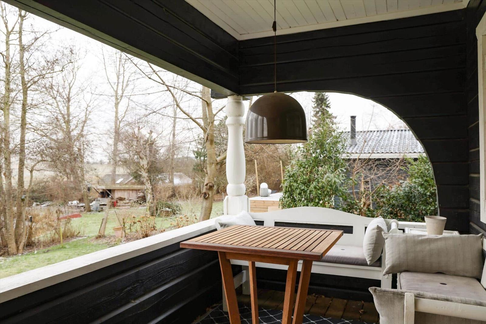 Veranda mit Tisch, Sitzbank und Hängelampe. Blick auf Garten und Bäume.