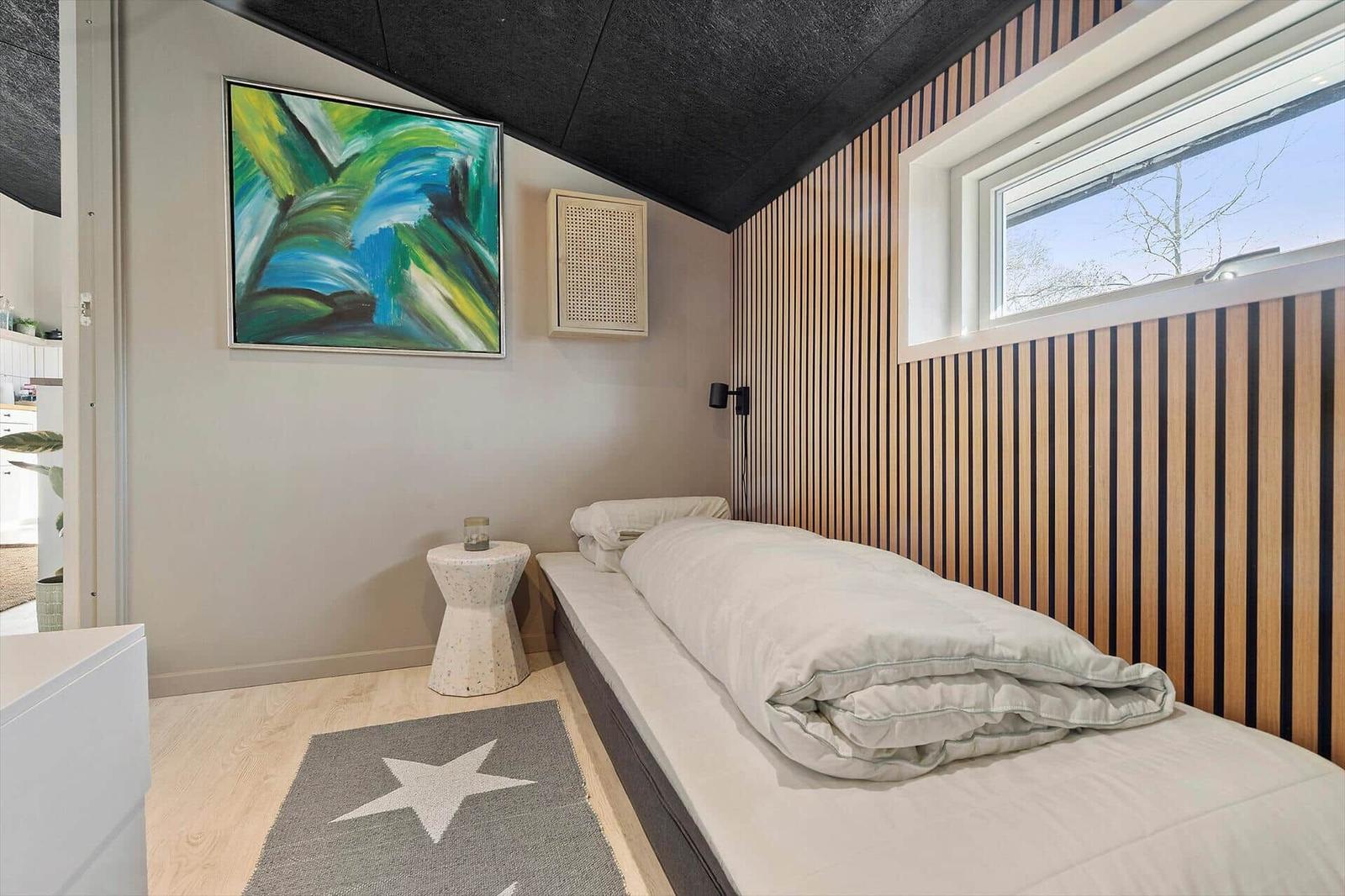 Schlafzimmer mit Bett, Wandbild und Holzverkleidung. Fenster und Bodenbelag sichtbar.