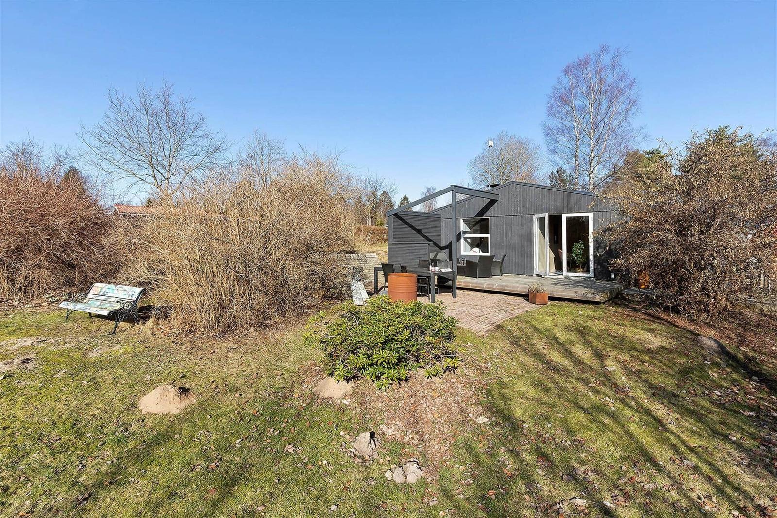 Moderne Hütte mit Terrasse, Garten und Sitzbank. Umgeben von Bäumen und Sträuchern.