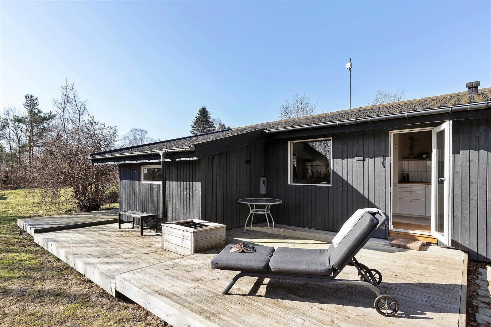 Holzterrasse mit Liegestuhl, Tisch und Grillbereich vor einem Haus mit offener Tür.