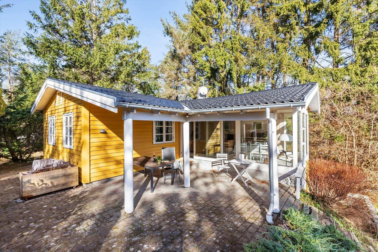Gelbes Holzhaus mit überdachtem Patio und Gartenbereich. Umgeben von Bäumen.