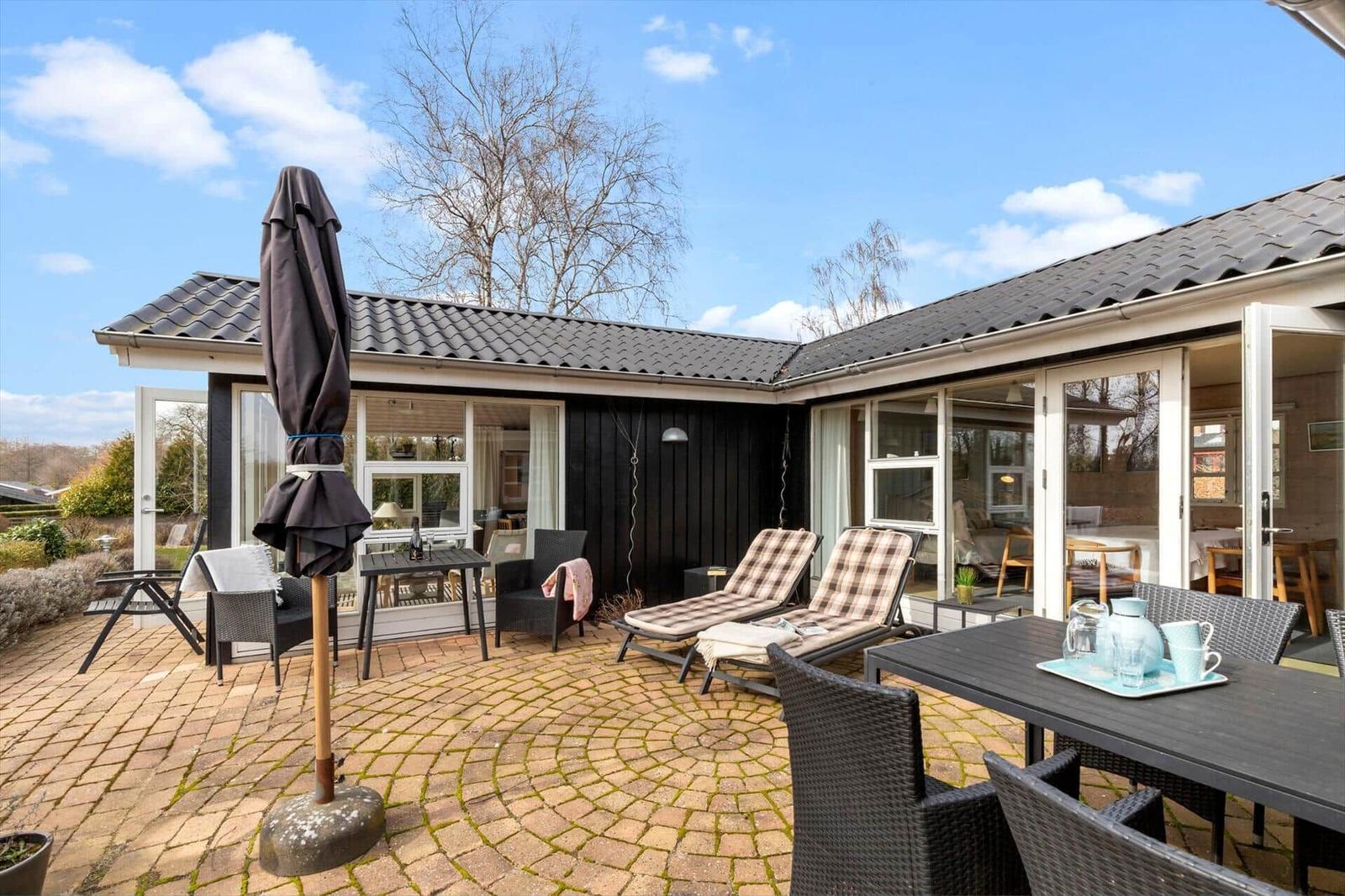 Terrasse mit Liegestühlen, Tisch und Sonnenschirm. Haus mit Glasfronten und dunklem Holz.