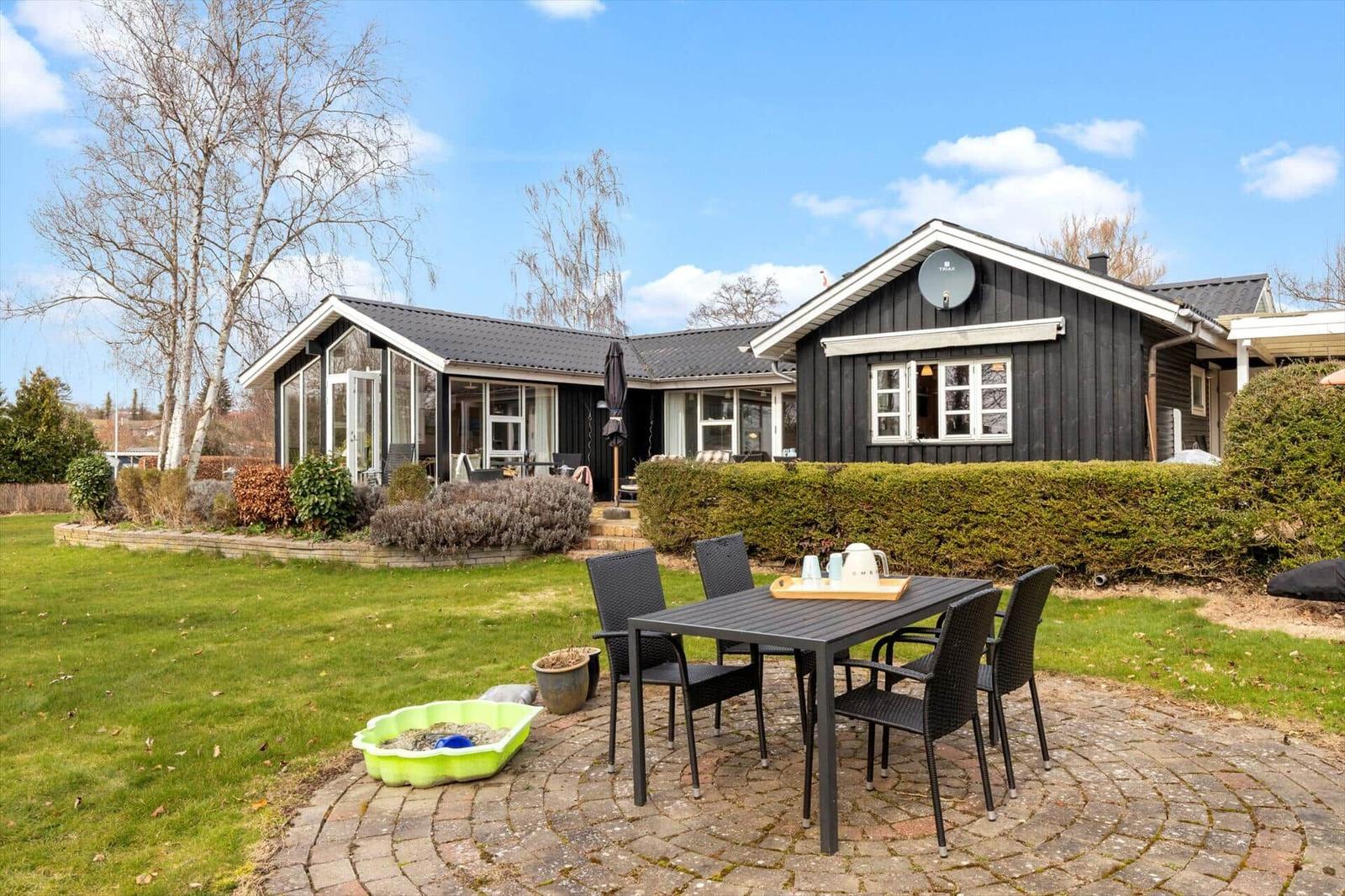 Schwarzes Holzhaus mit Terrasse und Garten. Außentisch mit Stühlen und Sandkasten.