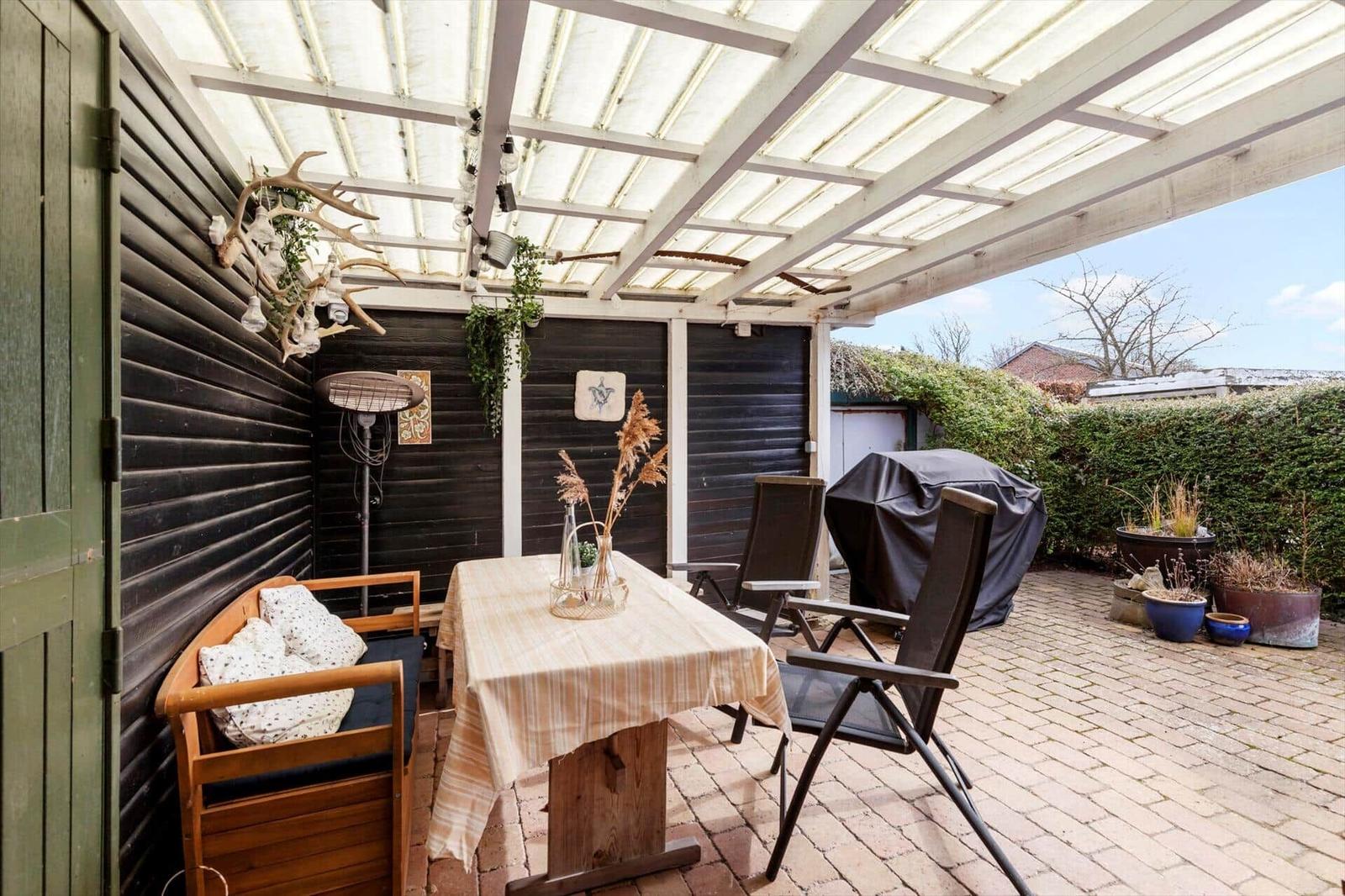 Terrasse mit Tisch, Stühlen und Grill unter Glasdach. Wand mit Dekoration und Pflanzen.