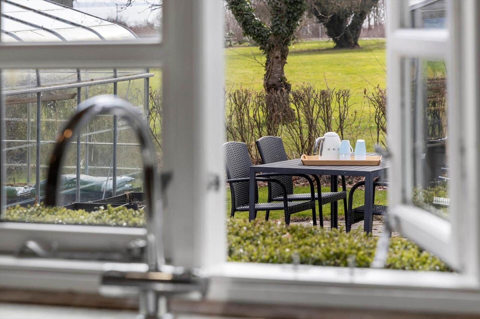Durch ein Fenster blickt man auf eine Terrasse mit Tisch, Stühlen und einer Teekanne.