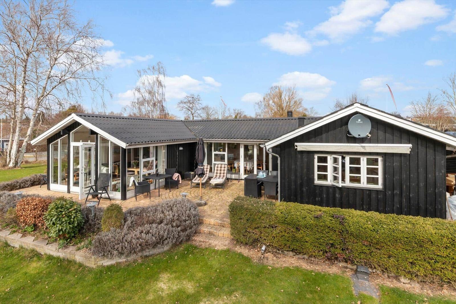 Schwarzes Holzhaus mit Terrasse und Garten. Satellitenschüssel und Fenster sichtbar.