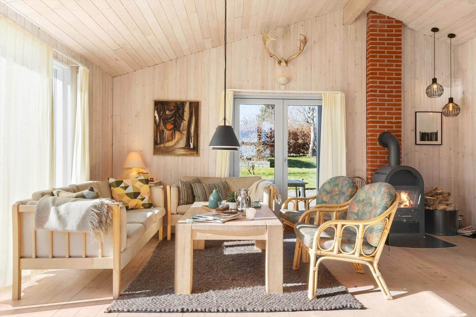 Wohnzimmer mit Holzwänden, Kamin und Sitzgelegenheiten. Fenster zur Außenterrasse.