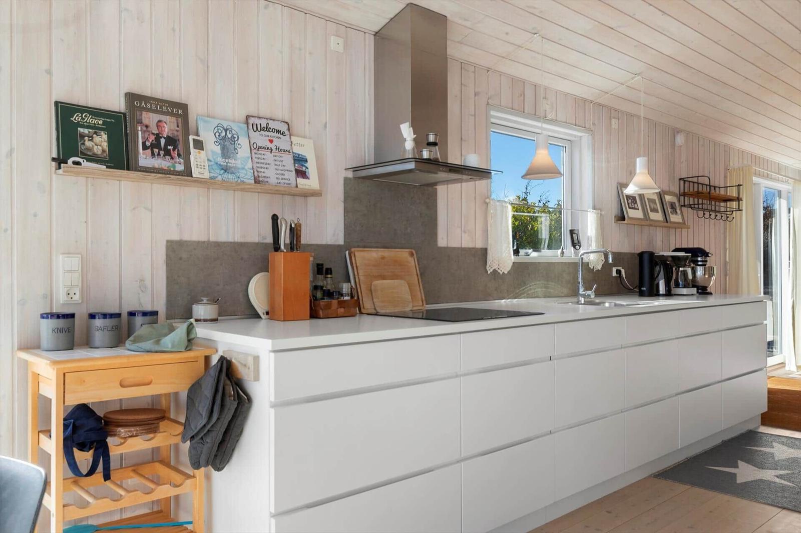 Moderne Küche mit weißen Schränken, Holzwand und Fenster. Arbeitsplatte, Spüle und Küchengeräte sichtbar.