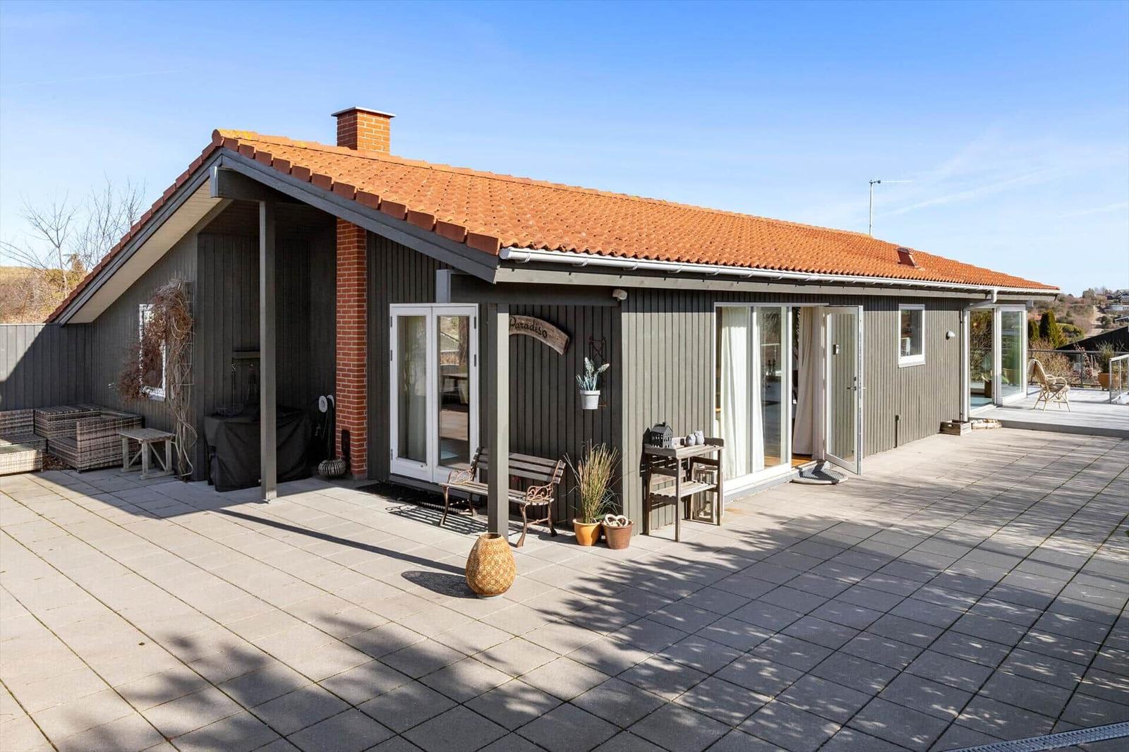 Ein Haus mit Terrasse, Glasfronten und rotem Ziegeldach. Außenbereich mit Möbeln und Pflanzen.