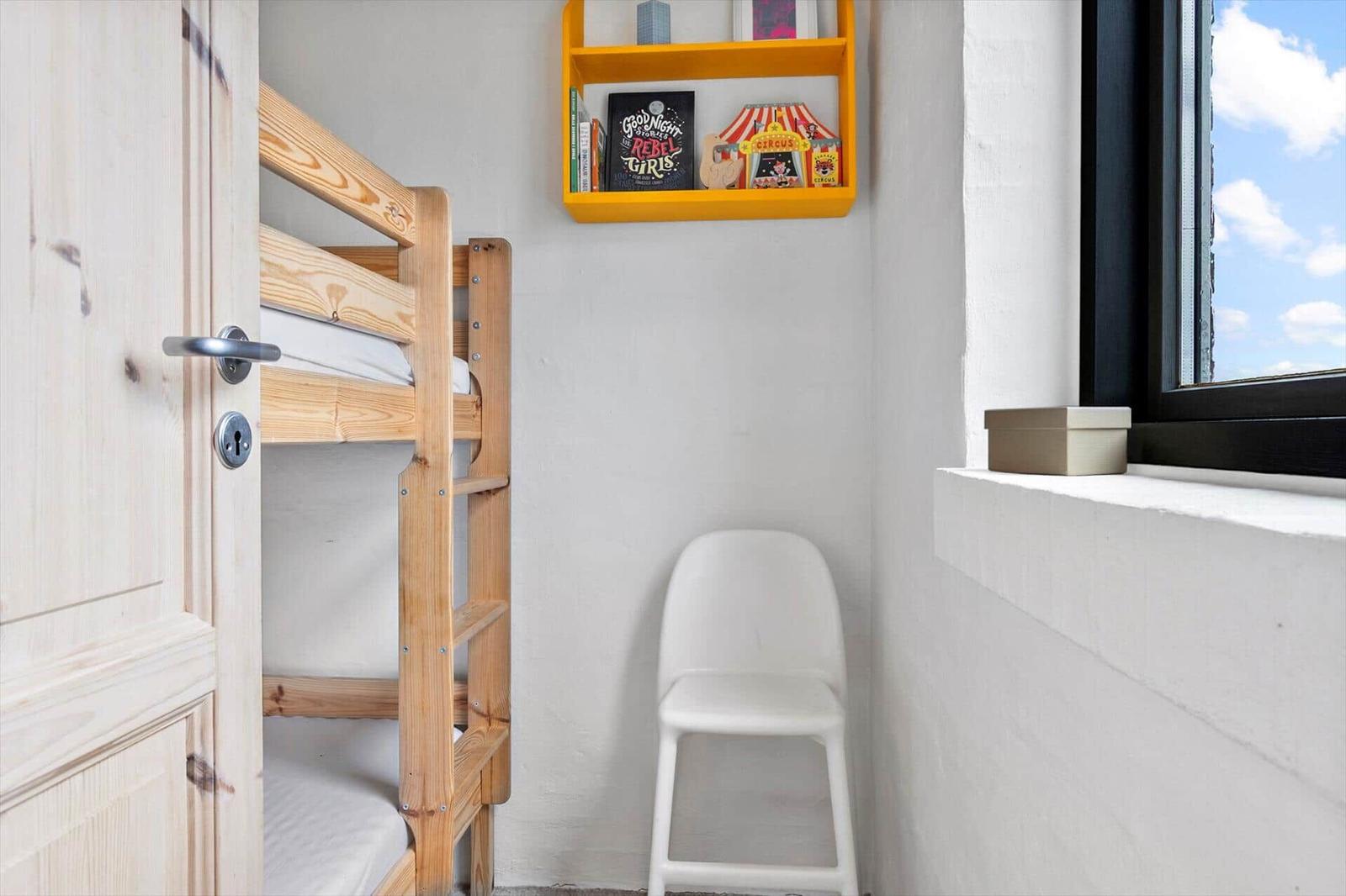 Kinderzimmer mit Holz-Etagenbett, weißer Stuhl und gelbem Regal an der Wand.