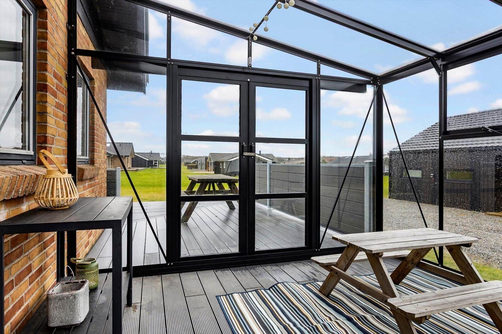 Terrasse mit Glasdach, Holztisch und Sitzmatte. Blick auf Garten und Nachbarhäuser.