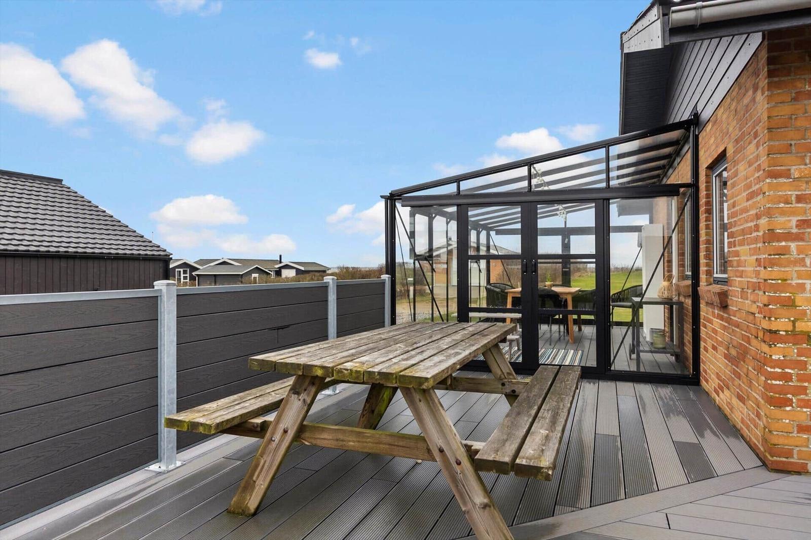 Terrasse mit Holzpicknicktisch und Glasüberdachung neben Backsteinhaus.