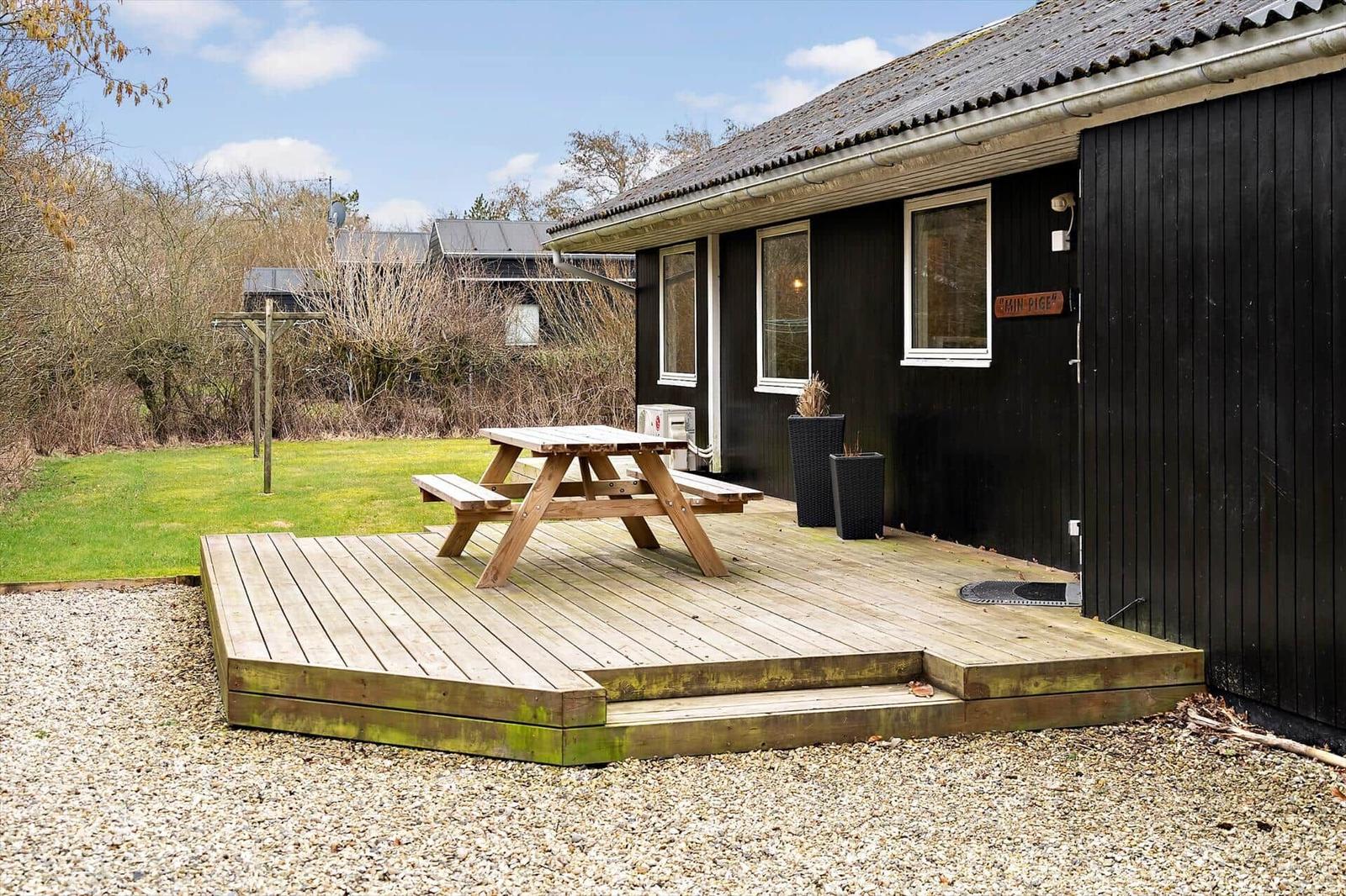 Schwarzes Haus mit Holzterrasse und Picknicktisch. Grasfläche und Bäume im Hintergrund.