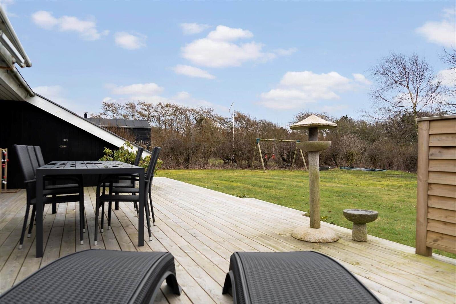 Terrasse mit Tisch, Stühlen und Liegestühlen. Grünfläche mit Spielgeräten und Steinobjekten.